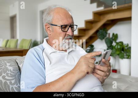 Älterer Mann mit Brille, der auf einer Couch sitzt und sich konzentriert, während er sein Smartphone in einem hellen, modernen Wohnzimmer benutzt. Stockfoto