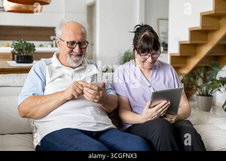 Glückliches älteres Paar, das zu Hause auf einer Couch sitzt und lächelt, während es Smartphone und Tablet für Kommunikation, Surfen und Freizeit in einem modernen Leben verwendet Stockfoto