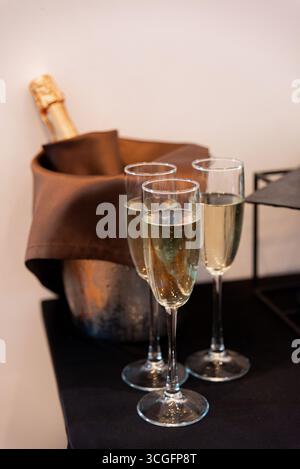 Drei Sektgläser gefüllt und bereit für eine Feier. Eine gekühlte Flasche Champagner befindet sich in einem Eiskübel, was die festliche Atmosphäre unterstreicht Stockfoto