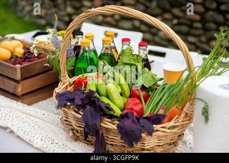 Ein Korbkorb voller lebendiger Gemüsesorten sitzt auf einem Tisch im Freien, begleitet von verschiedenen farbenfrohen Getränken in Flaschen. Perfekt für ein Gartentreffen Stockfoto