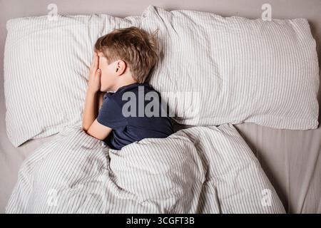 Das Kind hatte Angst vor dem Schlafengehen. Nachtschrecken bei einem Kind. Der Junge bedeckt sein Gesicht mit seinen Händen in Angst. Albträume und Angst vor der Dunkelheit. Stockfoto