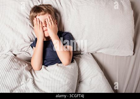 Das Kind hatte Angst vor dem Schlafengehen. Nachtschrecken bei einem Kind. Der Junge bedeckt sein Gesicht mit seinen Händen in Angst. Albträume und Angst vor der Dunkelheit. Stockfoto