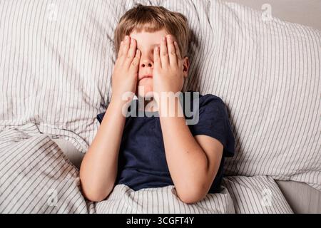 Das Kind hatte Angst vor dem Schlafengehen. Nachtschrecken bei einem Kind. Der Junge bedeckt sein Gesicht mit seinen Händen in Angst. Albträume und Angst vor der Dunkelheit. Stockfoto