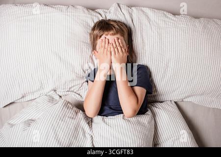 Das Kind hatte Angst vor dem Schlafengehen. Nachtschrecken bei einem Kind. Der Junge bedeckt sein Gesicht mit seinen Händen in Angst. Albträume und Angst vor der Dunkelheit. Stockfoto