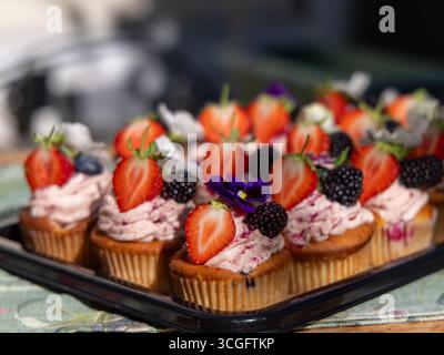 Tablett mit Cupcakes mit rosa Zuckerguss, belegt mit Erdbeeren, Brombeeren, Heidelbeeren und essbaren Blumen. Peggy Porschen Sommerrezept. 🔍 Stockfoto