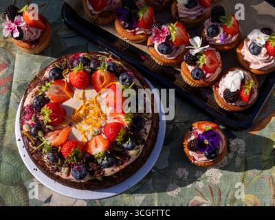 Tim Spectors Zoe-freundlicher Karottenkuchen mit Peggy Porschen Sommerbeeren-Cupcakes, dekoriert mit Früchten, Blumen und Nieseln auf floralem Tischtuch Stockfoto