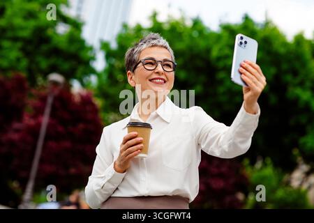Selbstbewusste, reife Geschäftsfrau macht ein Selfie im Freien mit Kaffee in der Hand in einem urbanen Park Stockfoto