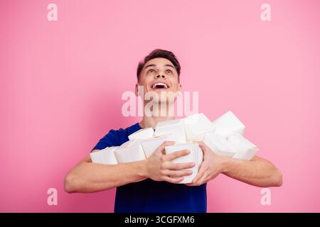 Fröhlicher junger Mann in einem blauen Hemd, der mehrere Take-Out-Behälter hält, während er vor einem leuchtend rosa Hintergrund lächelt Stockfoto