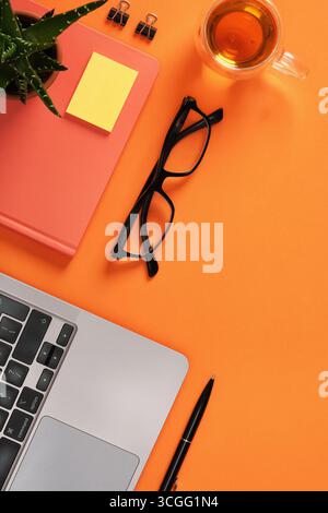 Laptop mit Notebook, Brille, Stift und Tasse Tee auf orangefarbenem Schreibtisch. Büroarbeitsplatz mit Copyspace Stockfoto