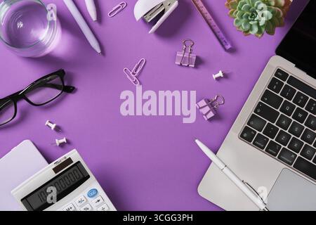Schreibtisch mit Laptop, Stift, Hefter, Brille, Büroklammer, und Taschenrechner auf violettem Hintergrund. Platzieren für Ihren Text. Stockfoto