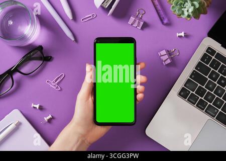 Handgriff Smartphone mit grünem Bildschirm. Laptop, Schreibwaren und Glas Wasser auf violettem Schreibtisch. Platz für Screenshot. Stockfoto
