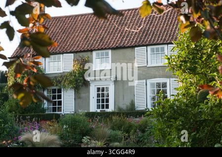Ein Landhaus zeigt eine graue Außenseite und weiße Fensterläden, eingebettet zwischen bunten Blumen und grünem Laub im Nachmittagslicht. Stockfoto