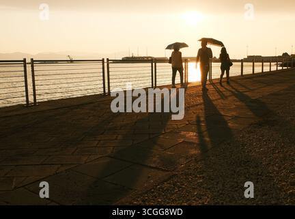 Silhouettenfiguren, die bei einem ruhigen Sonnenuntergang an einer Uferpromenade entlang laufen, mit Sonnenschirmen in der Hand, die lange Schatten auf den Bürgersteig werfen. Stockfoto