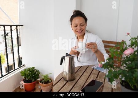 Eine glückliche junge Frau in einem weißen Gewand genießt ihren Morgenkaffee auf einem hübschen Balkon, der mit Pflanzen und Blumen dekoriert ist. Die Szene strahlt Entspannung und W aus Stockfoto