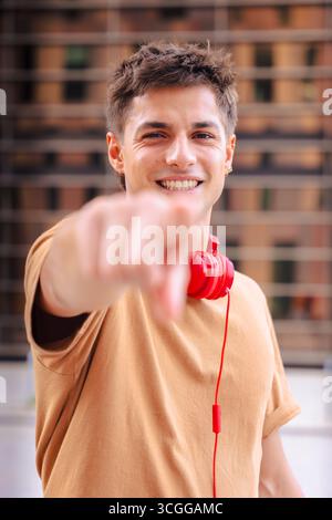 Ein lächelnder junger Mann, der auf die Kamera zeigt und draußen rote Kopfhörer trägt. Vertikal Stockfoto