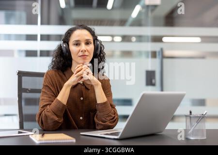 Konzentrierte Frau im Geschäftsumfeld mit Kopfhörern, auf dem Schreibtisch mit Laptop und Notebook. Stockfoto