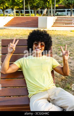 Junger Mann, der ein Friedenszeichen zeigt, während er sich auf der Bank im Park entspannt Stockfoto