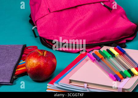 Rosafarbener Rucksack, Notizbücher, Stifte, Bleistiftetui und Apfel auf türkisfarbenem Hintergrund für den Schulbesuch Stockfoto