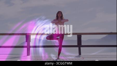 Ausbalancierender Yogapraktiker, der auf einem Holzsteg am Wasser mit digitalem Lichtwirbel eine Baumposition ausführt Stockfoto