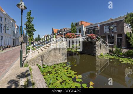 Eine charmante Steinbrücke über einen Kanal in Gouda, die mit Blumenkörben geschmückt ist, überquert einen Kanal mit Lilienpads, eingerahmt von historischen niederländischen Gebäuden unter einem c Stockfoto