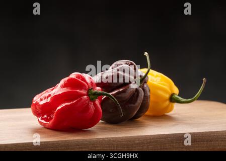 Drei frische Habanero-Paprika in den Farben Rot, Schokoladenbraun und Gelb auf einem hölzernen Küchenbrett vor dunklem Hintergrund Stockfoto
