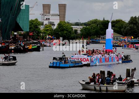 Schweben Sie mit aufblasbarer Spritze und die kostümierten Teilnehmer fahren auf der Canal Parade am Pride Amsterdam in Amsterdam, Niederlande, vorbei an jubelnden Menschenmassen. Stockfoto
