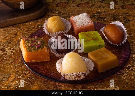 Indische Süßigkeiten in Pappbechern auf dekorativem Teller. Traditionelle Mithai flach auf goldenem Tuch für festliche Lebensmittelfotografie. Stockfoto