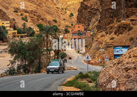 Mit dem Auto fahren Sie durch eine gewundene Bergstraße mit Palmen, felsigen Klippen und traditionellen marokkanischen Häusern in einem Wüstental Stockfoto