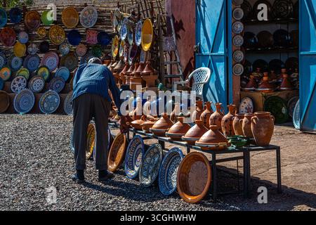 Ein einheimischer Mann arrangiert traditionelle marokkanische Tagine-Töpfe und bunte Keramikplatten auf einem Töpfermarkt im Freien in Marokko. Stockfoto
