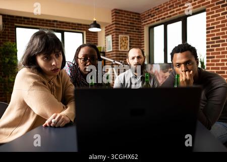 Eine Gruppe von Freunden sitzt um den Tisch in einer gemütlichen Wohnung und sieht Filme auf einem Laptop mit seriösem Aussehen an. Verschiedene Personen saßen drinnen und genossen ein entspanntes Wochenende bei Videoinhalten. Stockfoto