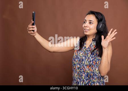 Entspannte indische Frau mit schnurlosen Kopfhörern, die das Handy hält, lächelt und winkt auf dem Bildschirm. Fröhliche weibliche Person, die Videoanrufe mit der Familie hat und vor braunem Hintergrund steht. Stockfoto