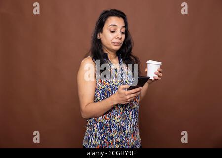Indianerin steht vor braunem Hintergrund, hält ihren Kaffee und benutzt ein Smartphone. Das weibliche Model hält beiläufig einen Take-away-Becher und blättert durch ihr mobiles Gerät, um sich online zu unterhalten. Stockfoto