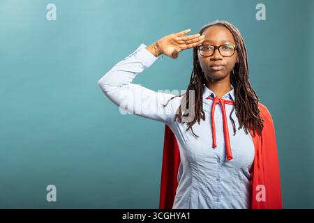 Ernste schwarze Heldin steht und respektvollen militärischen Gruß gibt. Die junge afroamerikanische Frau in rotem cape und blauem Hemd schlägt kraftvolle Posen und strahlt Vertrauen im Studio aus. Stockfoto