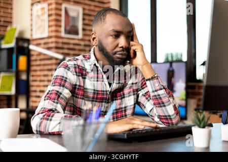Ein bärtiger männlicher Manager arbeitet von zu Hause aus und sieht frustriert und gelangweilt aus, während er auf dem Computer Projektaktualisierungen überprüft. Selbstständiger Schwarzer sitzt im Wohnzimmer, überfordert von digitalen Aufgaben. Stockfoto