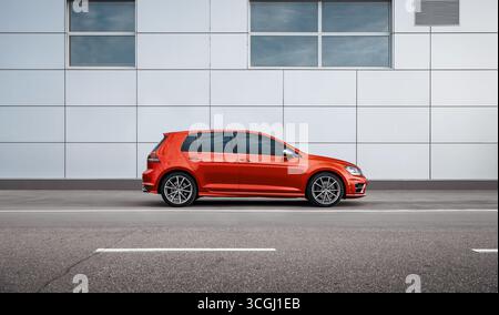 Rotes Auto auf der Straße im Industrieviertel. Seitenansicht des deutschen Volkswagen Golf Fließhecks, der in einer Stadt in der Nähe einer grauen Mauer eines modernen Gebäudes geparkt ist Stockfoto