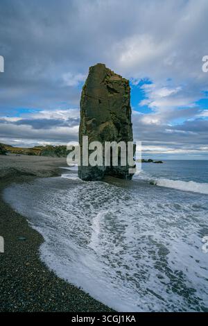 Am schwarzen Sandstrand Islands erhebt sich ein gewaltiger Meeresstapel, während die Wellen unter dramatischen Abendwolken gegen seine Basis krachen Stockfoto