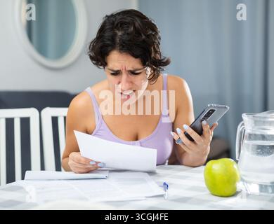 Schockierte junge Frau, die Dokumente ansieht und Smartphone in der Hand hält Stockfoto