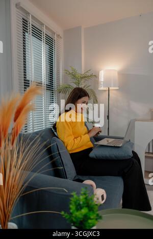Junge asiatische Frau in gelbem Sweatshirt lächelt, während sie etwas auf ihrem Laptop sieht und eine Tasse Kaffee hält. Sie sitzt bequem auf einem Sofa in A Stockfoto