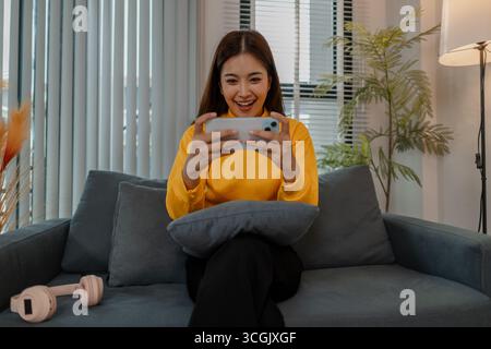 Junge asiatische Frau in gelbem Sweatshirt, die zu Hause auf dem Sofa sitzt und sich intensiv auf das Spielen von mobilen Spielen auf dem Smartphone konzentriert. Entspannender Innenstil mit Stockfoto