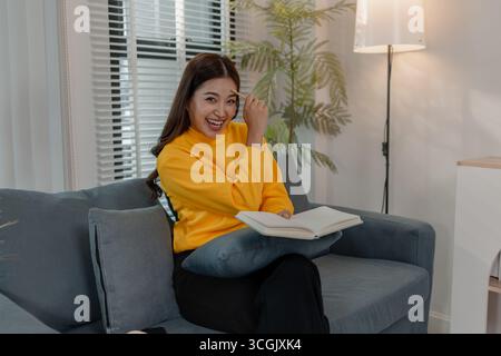 Fröhliche asiatische Frau in gelbem Pullover sitzt auf dem Sofa, lacht und hält einen Bleistift an den Kopf mit einem offenen Notizbuch auf dem Schoß. Konzept von inspira Stockfoto