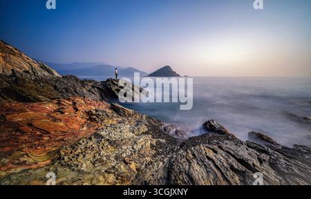 Eine Einzelfigur steht auf zerklüfteten Felsen mit Blick auf das Meer, während weiche Wellen gegen das Ufer krachen. Stockfoto