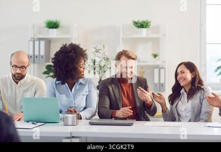 Fröhlich lächelnde Mitarbeiter aus verschiedenen Büros treffen sich im Konferenzraum beim Brainstorming am Schreibtisch, diskutieren gemeinsam Stockfoto