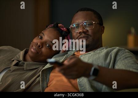 Porträt der jungen Schwarzen Frau und des jungen Schwarzen Mannes, die eng zusammen auf dem Sofa sitzen, Mann mit Fernbedienung und Blick in die Kamera, Frau, die Kopf auf der Schulter des Mannes liegt Stockfoto
