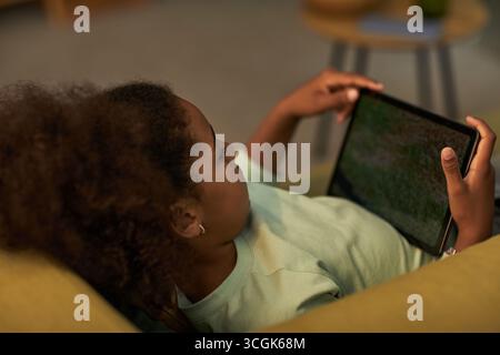 Schwarzes Mädchen, das auf dem Sofa liegt und ein digitales Tablet hält, mit beiden Händen auf dem Bildschirm schaut, sich zu Hause entspannt, lockiges Haar sichtbar, legere Umgebung, Technologie wird von Kindern genutzt Stockfoto
