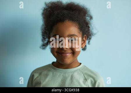 Porträt eines Schwarzen Mädchens, das in die Kamera lächelt, vor blauem Hintergrund steht, natürliches lockiges Haar zeigt und kleine Creolen trägt, die das Glück der Kindheit repräsentieren Stockfoto