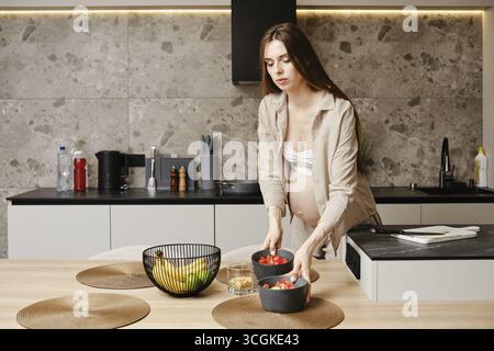 Eine schwangere Frau in einer modernen Küche legt sorgfältig frischen Salat in Schüsseln und bereitet sich auf eine gesunde Mahlzeit vor Stockfoto