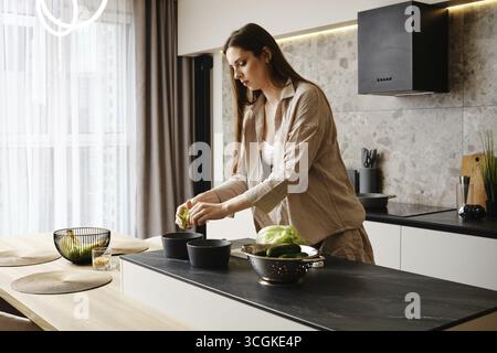 Eine Frau mit langen Haaren schneidet Gemüse in einer modernen Küche. Sie konzentriert sich auf ihre Aufgabe, während sie auf einer Steinplatte steht Stockfoto