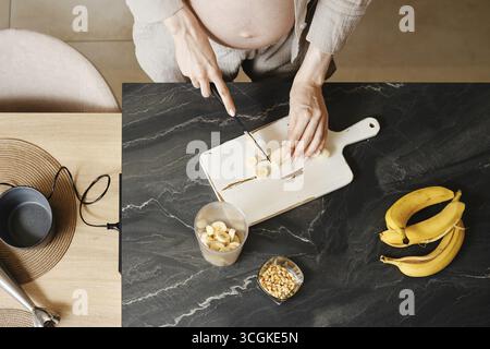 Eine Person mit einem sichtbaren Babypuffer schneidet Bananen auf einem hölzernen Schneidebrett in einer modernen Küche. Frische Bananen und geschnittene Stücke sind ordentlich angeordnet Stockfoto
