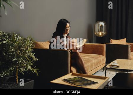 Eine Frau sitzt bequem auf einer modernen Couch und ist in einer stilvollen Lounge im Innenbereich vertieft, ein Buch zu lesen Stockfoto