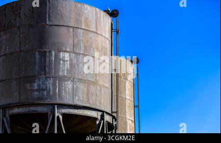 Zwei große, verrostete Metalltanks mit blauem Himmel im Hintergrund. Die Tanks sind alt und haben ein verwittertes Aussehen Stockfoto